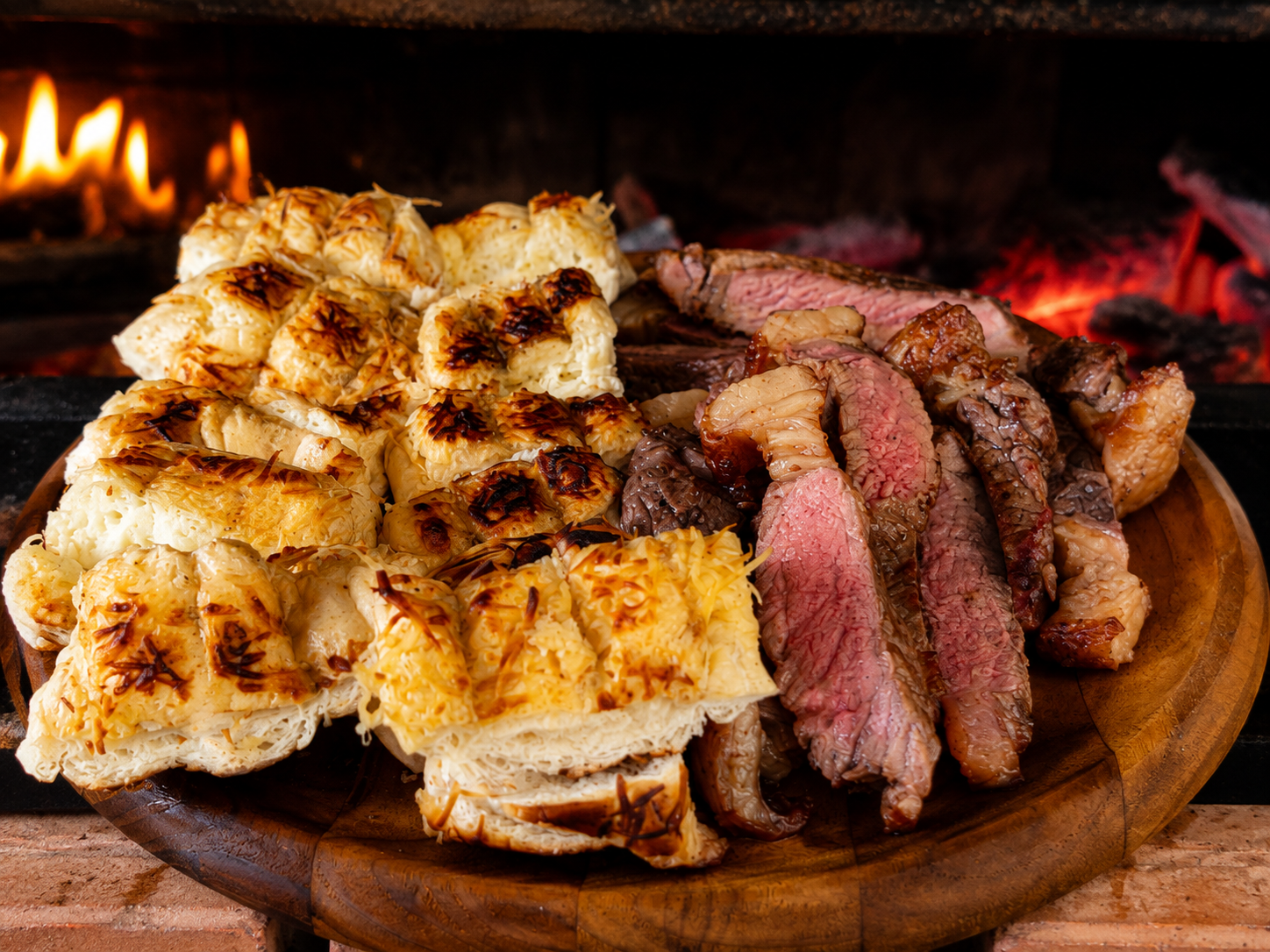 Foto de pães de alho e bife ancho numa tábua com brasas ao fundo